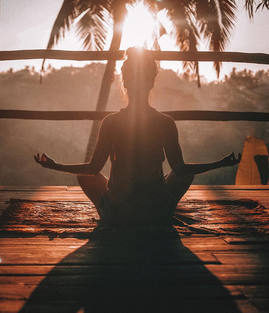 Person meditating in a cross-legged position on a wooden deck at sunset, with palm trees in the background.